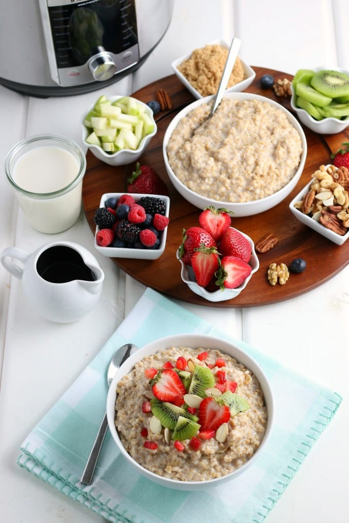 A lazy susan with a large bowl of oatmeal surrounded by smaller bowls of fresh toppings - berries, strawberries, nuts, kiwi and brown sugar. Milk and maple syrup are in small containers off to the side. A prepared bowl of oatmeal is in the foreground with a cloth napkin and spoon.