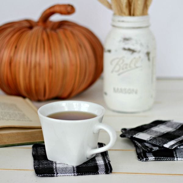 Image shows a coffee cup filled with coffee sitting atop a flannel homemade coaster. A mason jar, book, and pumpkin sit in the background.