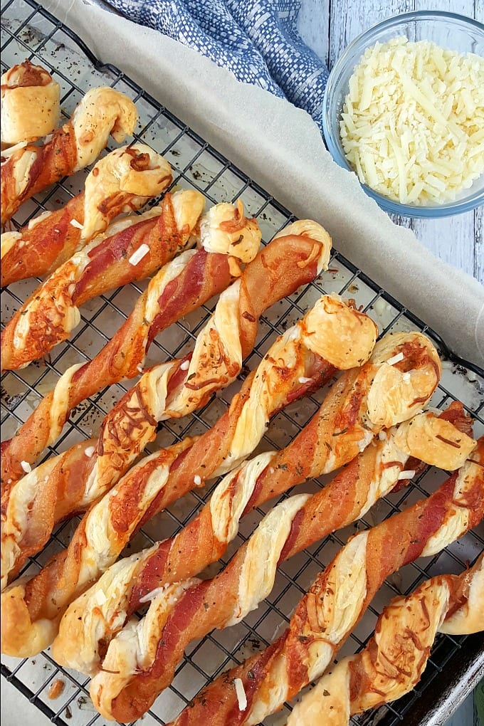 Bacon puff pastry twists lined up on a sheet pan on top of parchment paper and a rack. A small glass bowl full of Parmesan cheese sits next to the sheet pan.
