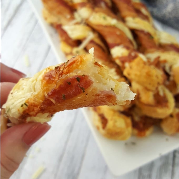Bacon puff pastry twists sit on top of a white plate. In the foreground is a torn twist being held by a hand.