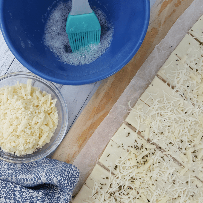 How to make bacon puff pastry twists: bowl with egg wash, bowl of cheese, and puff pastry on a cutting board.