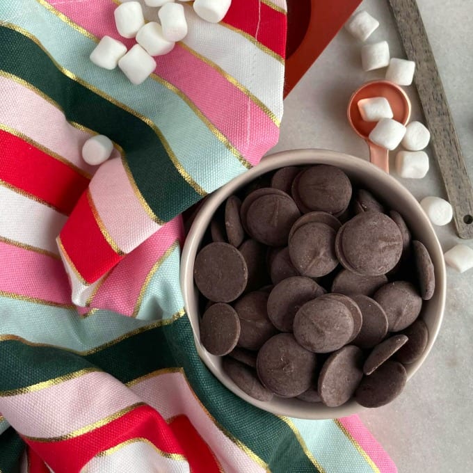 Melting chocolate wafers in a small ramekin surrounded by mini marshmallows and a cloth napkin.