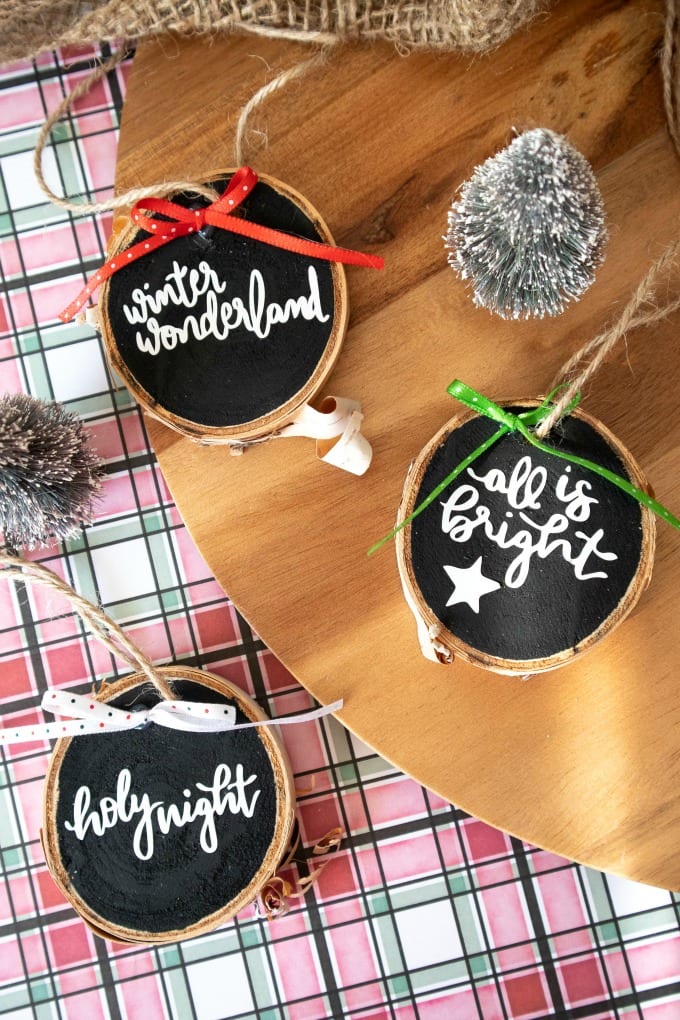A birds-eye-view of wood slice Christmas ornaments, black with white text. Sitting on red and green plaid background. 