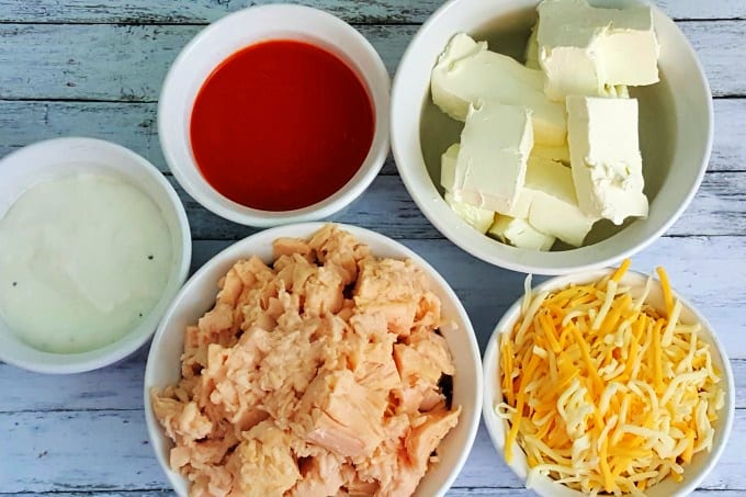 close up of buffalo chicken dip filling ingredients in small white bowls include ranch dressing, buffalo wing sauce, cream cheese, shredded chicken and shredded cheese.