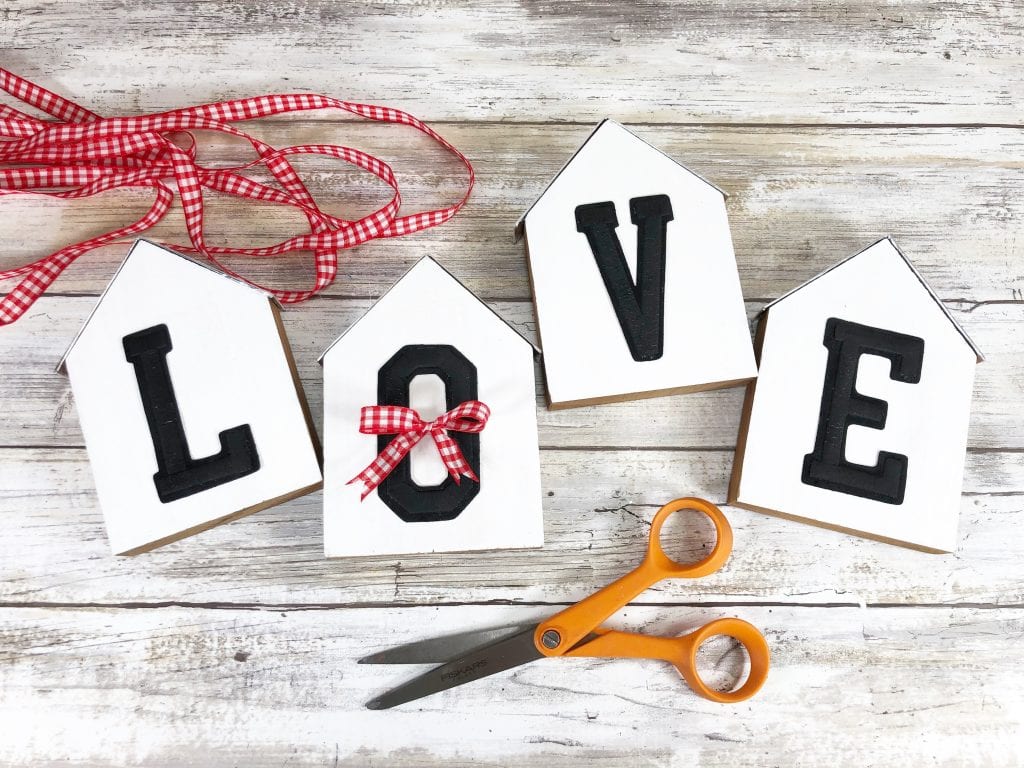 Decorative Wooden Houses - DIY Tutorial - four wooden blocks in the shape of houses with the letter L-O-V-E on them on a white background with red and white gingham ribbon and a pair of orange handled scissors