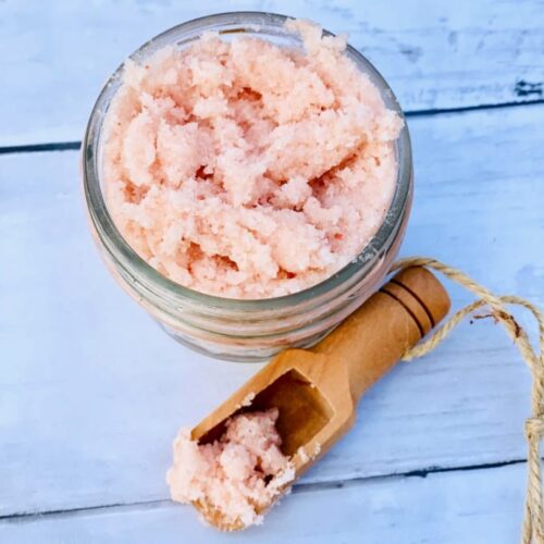 Top down image of pink Himalayan salt scrub in a glass jar with a wooden scoop nearby.