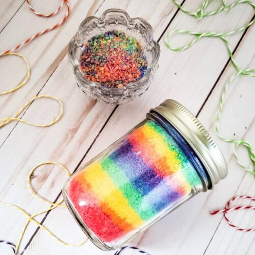 Rainbow bath salts in a glass mason jar next to a small bowl filled with the same.