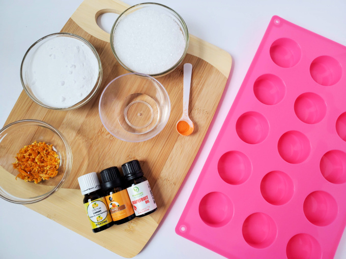 Image contains items you will need to make essential oil shower steamers - citrus oils, dried orange zest, water, baking soda, citric acid, mica powder and a silicone mold. 