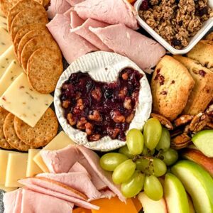 Baked brie on a platter with crackers, cheese slices, grabes and slices of ham.