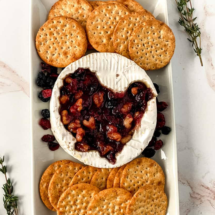 Top down view of baked brie, topped with cranberry sauce, on a white platter with dried cranberries and crackers. 