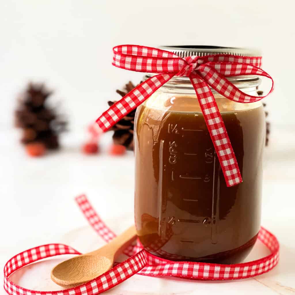 Homemade caramel in a glass jar with a red and white checked ribbon wrapped around the neck ready for gifting.
