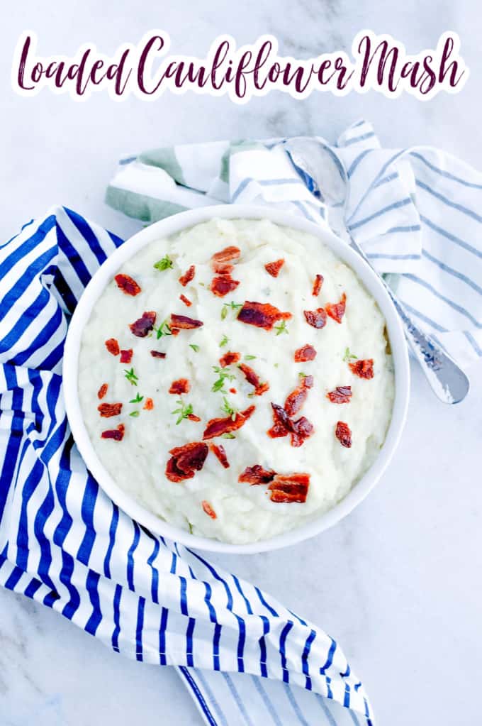 Top down image of loaded cauliflower mash in a white bowl, topped with bacon and fresh herbs. 