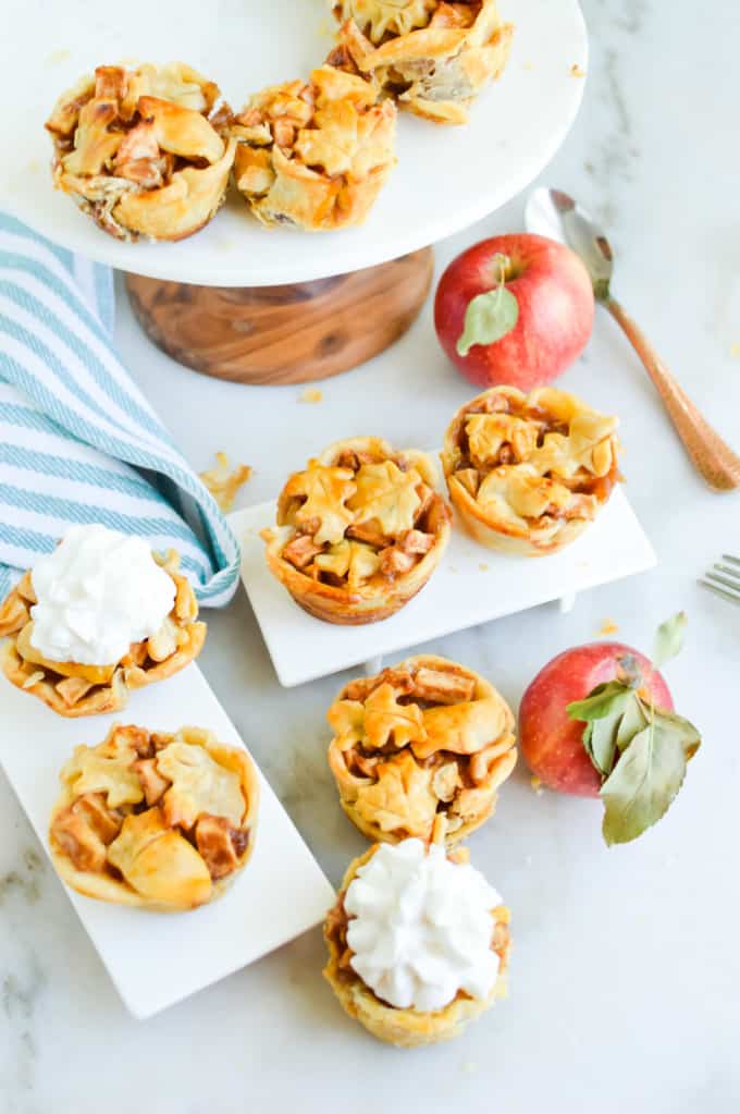 Mini Apple Pie Tarts on an assortment of white serving platters, surrounded by fresh apples.
