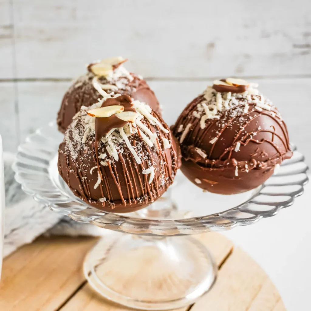 Three Almond Joy inspired hot chocolate bombs topped with coconut and almond slivers on a glass cake plate.