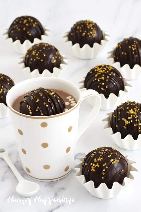 Boozy cocoa bombs topped with gold star sprinkles surround a white and gold polka-dotted mug filled with hot cocoa and mini marshmallows. 