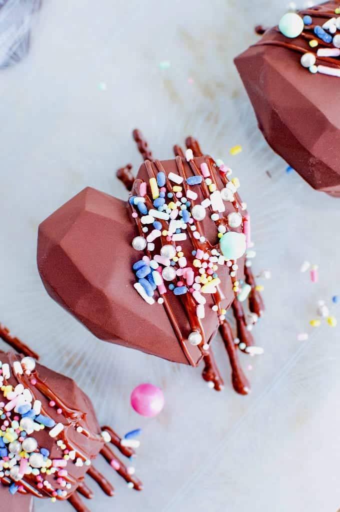 Heart shaped hot chocolate bombs covered in a chocolate drizzle and sprinkles.