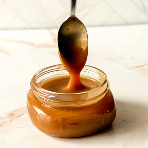 Close up image of a spoon pouring caramel sauce into a small glass jar.