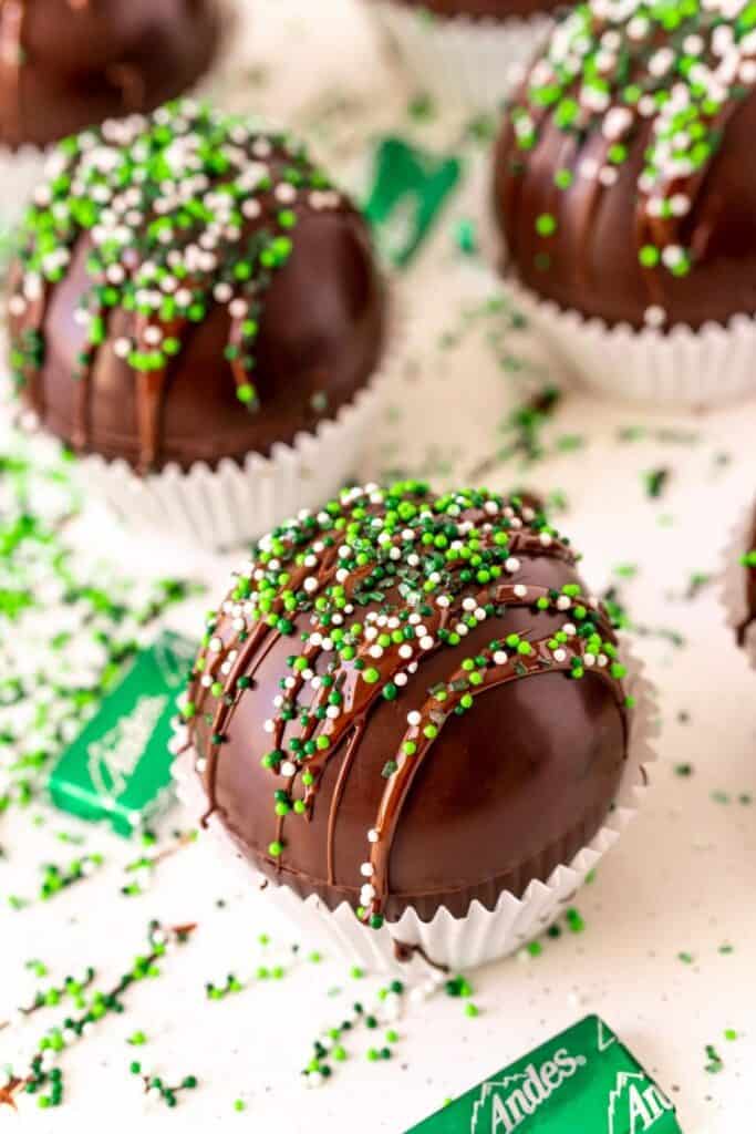 Close up image of a mint hot chocolate bomb in a white mini muffin tin liner, topped with a chocolate drizzle and green and white sprinkles. Andes mints surround it.