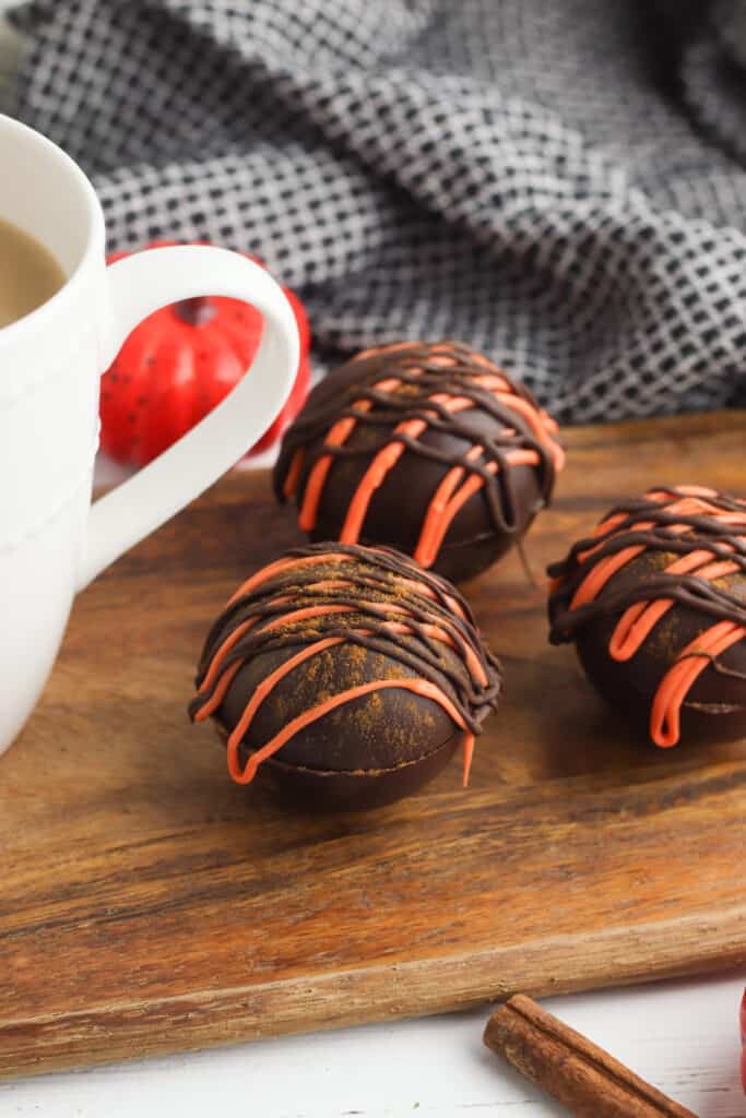 Close up image of cocoa bombs topped with an orange and brown chocolate drizzle, next to a white mug.