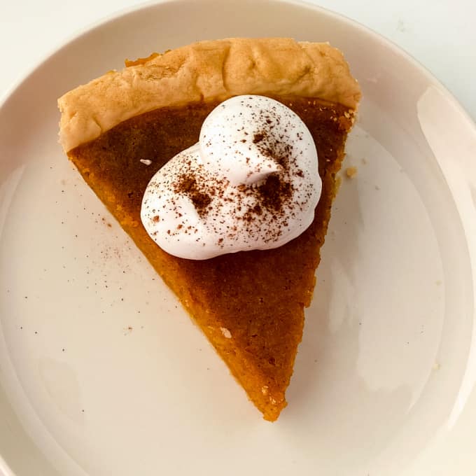 Image contains a slice of homemade sweet potato pie on a white plate, topped with whipped cream and cinnamon.
