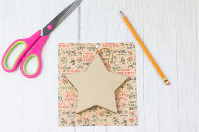 Star shaped, paper mache ornament sitting on top of a piece of scrapbook paper next to a pencil and scissors.