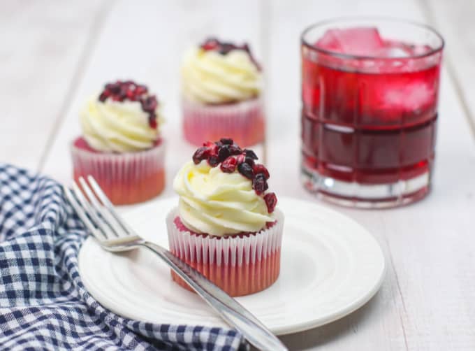 Pomegranate cupcake on a white plate with a fork, more cupcakes and a cocktail are behind it.