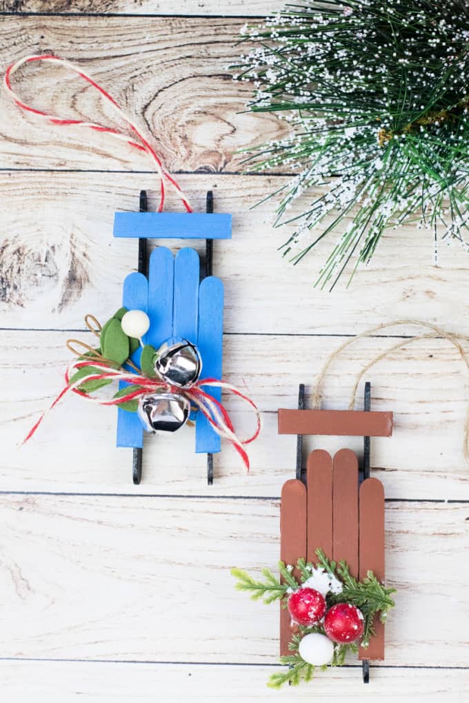 Two popsicle stick sled ornaments on a wood surface with Christmas greenery.