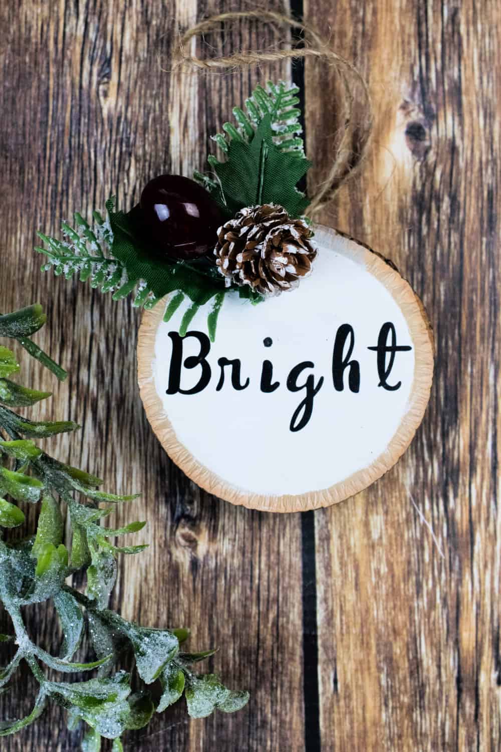 Close up image of a wood circle ornament painted white with the word "Bright" on it along with Christmas greenery.