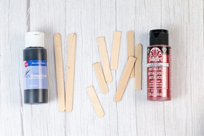 Black craft paint next to two popsicle sticks next to cut pieces of popsicle sticks and a bottle of metallic craft paint.