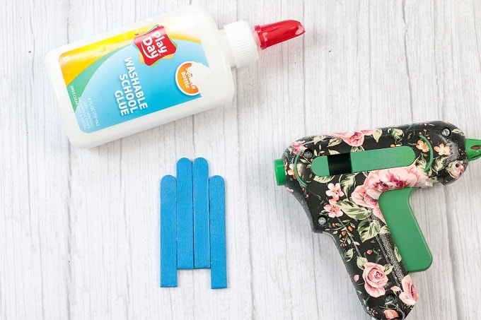 Image contains a bottle of glue, a hot glue gun, and 4 cut down popsicle sticks waiting to be glued together.