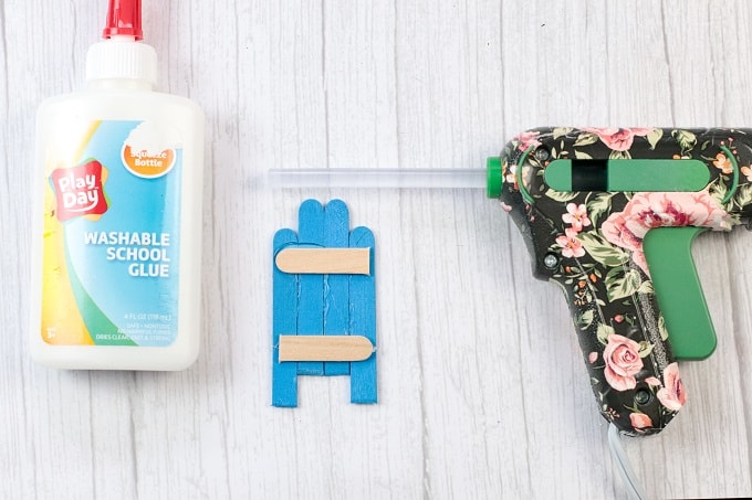 Image contains a bottle of glue, popsicle sticks being glued together to make a sled and a glue gun with a hot glue stick.