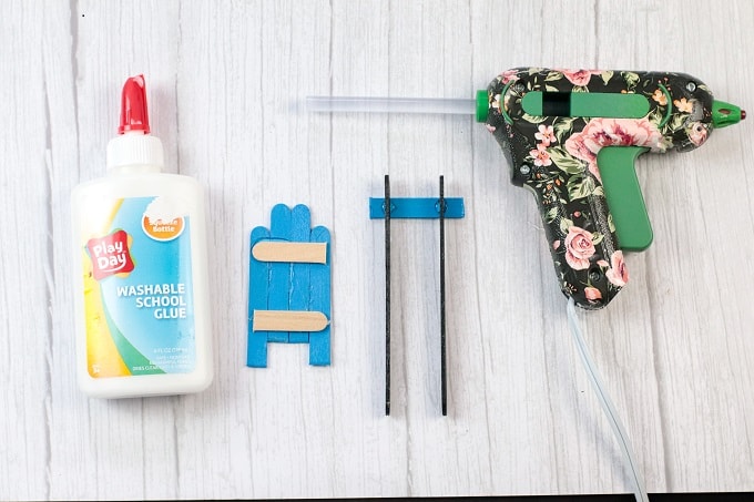 Image contains a bottle of glue, popsicle sticks that have been glued together to make pieces of a sled, and a hot glue gun.