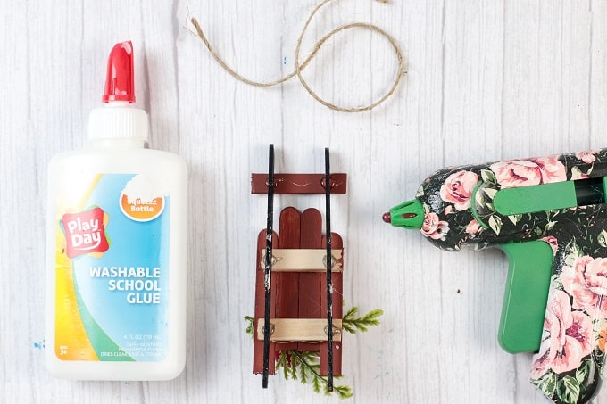 Image contains a bottle of glue, a popsicle stick ornament waiting for a twine loop to be glued on and a hot glue gun.