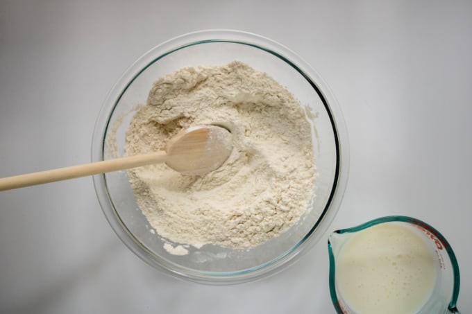 Wooden spoon in a mixing bowl full of dry ingredients next to a liquid measuring cup filled with buttermilk.