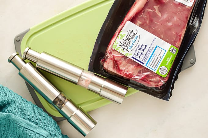 New York Strip Steak in a package from the grocery store on the counter with a cutting board and salt and pepper.