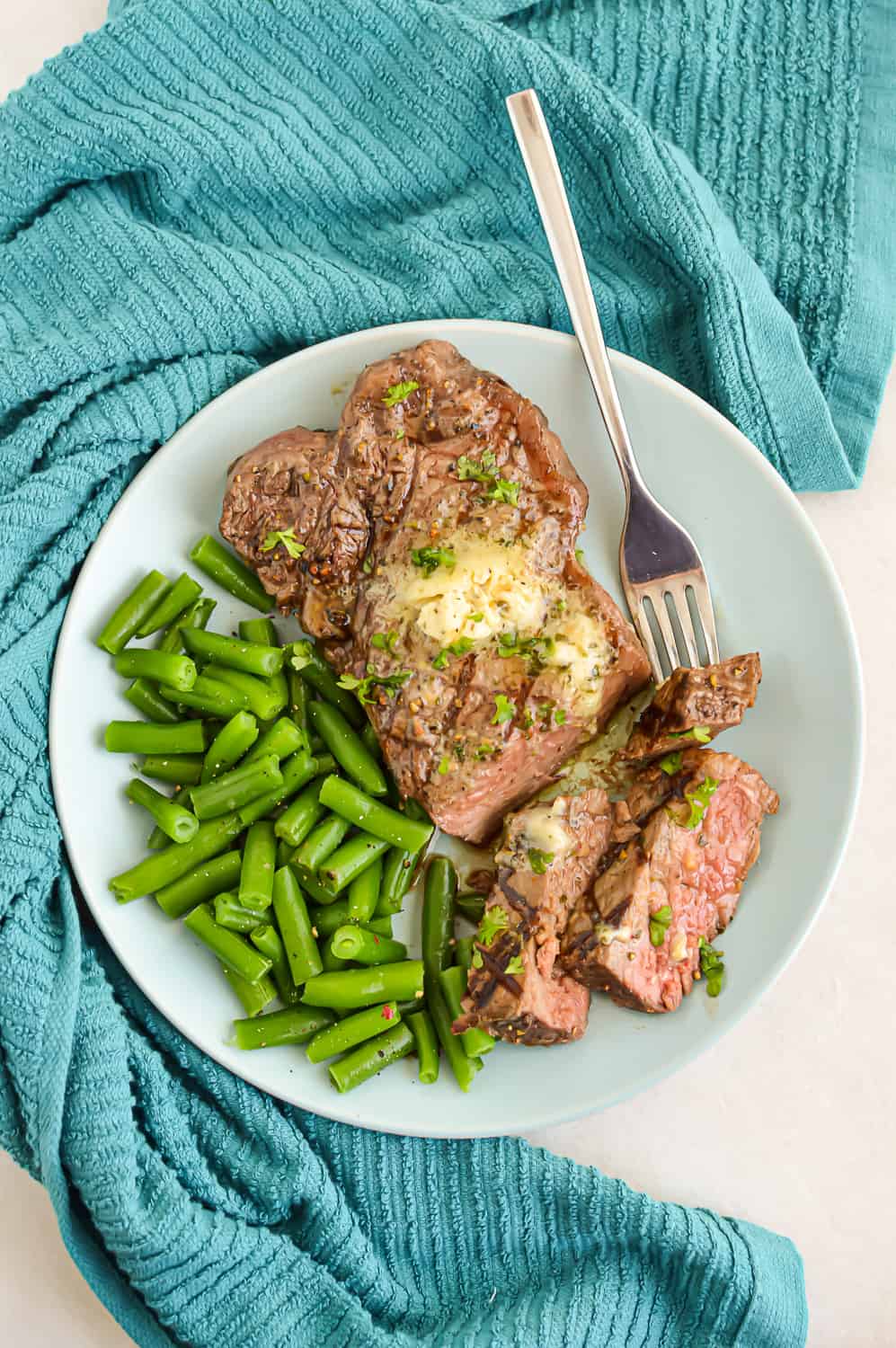 Vertical image of Broiled New York Strip Steak on a white plate with garlic herb butter and green beans.