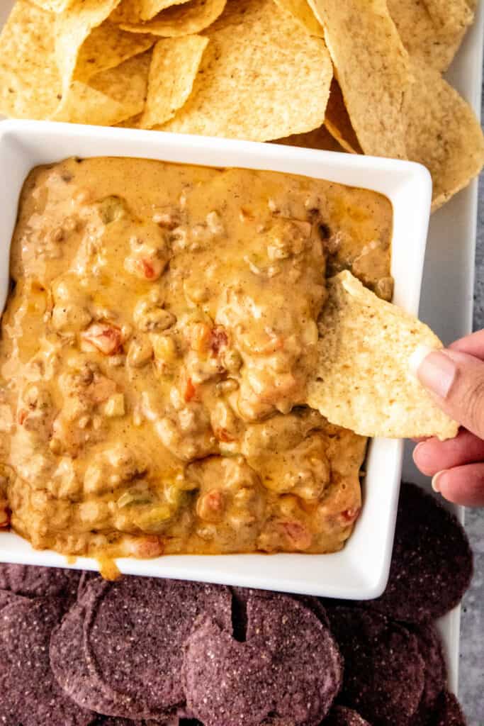 Hand holding a chip dipping into Queso Dip with meat in a square bowl with tortilla chips and blue corn chips.