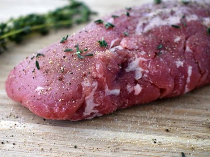 Close up of seasoned uncooked pork tenderloin on a cutting board.
