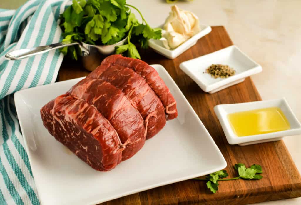 Petite sirloin roast ona white plate with olive oil, seasonings, garlic and parsley nearby.