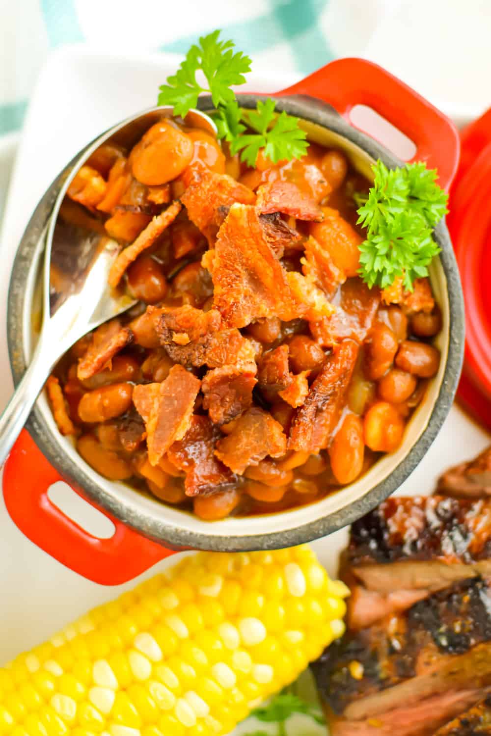 Close up of Cowboy Baked Beans topped with bacon in a small crock with a spoon next to corn on the cob.