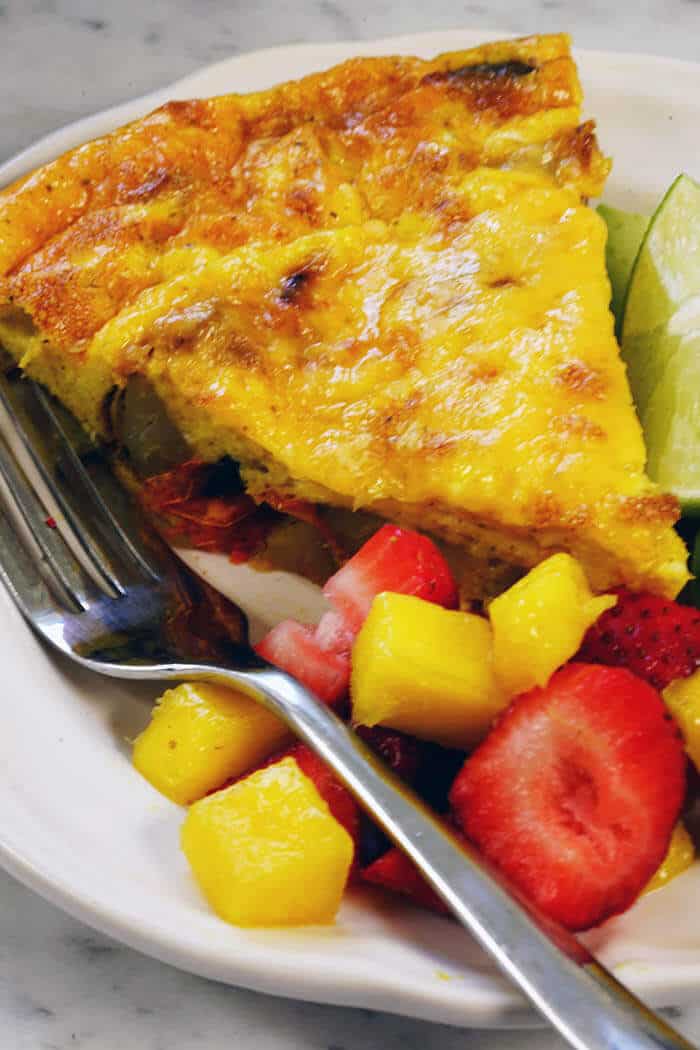 An easy frittata recipe pictured here. A white plate with a wedge of frittata served with avocado slices and fruit salad.