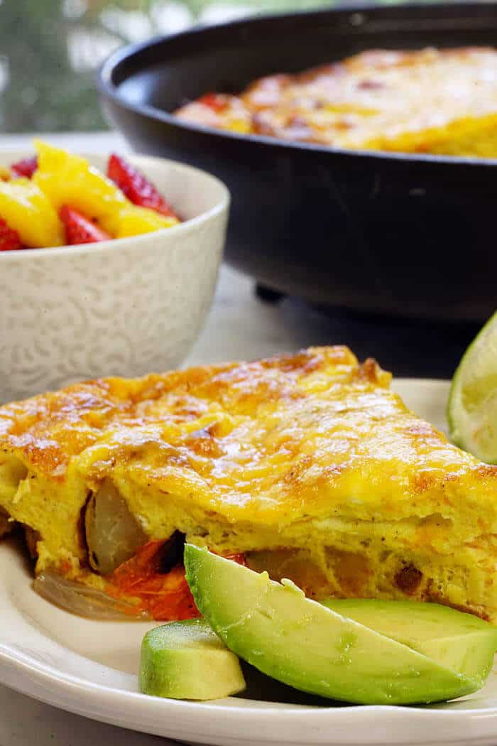 A white plate with a wedge of frittata served with a side of avocado and fresh fruit. This is a recipe for an easy frittata