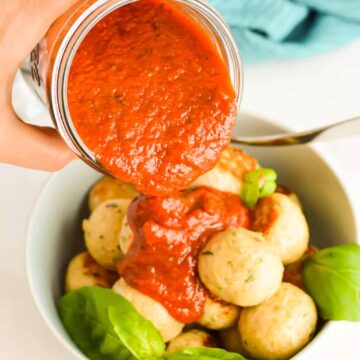 Hand pouring a jar of marinara sauce on top of Italian Chicken Meatballs.