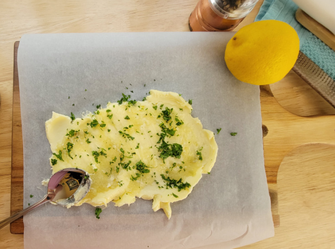 Butter, garlic, and herbs on a cutting board with a spoon and lemon.