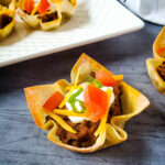 Taco Wonton Cups with tomatoes, sour cream, cheese, and green onions on a countertop.