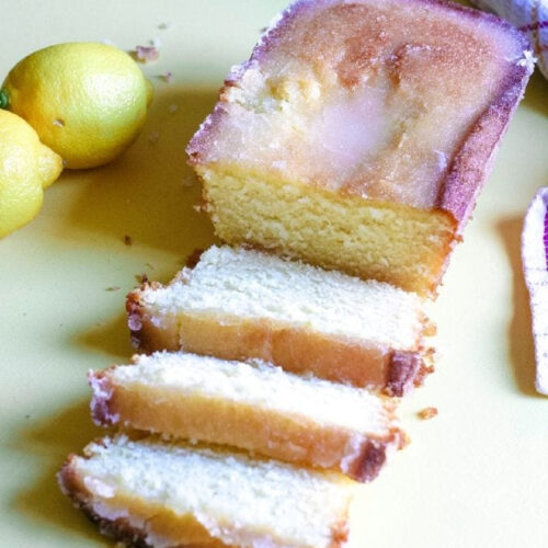A loaf of glazed Lemon Bread sliced next to fresh lemons.