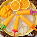 Orange Mango Pops in a bowl of ice next to fresh fruit.