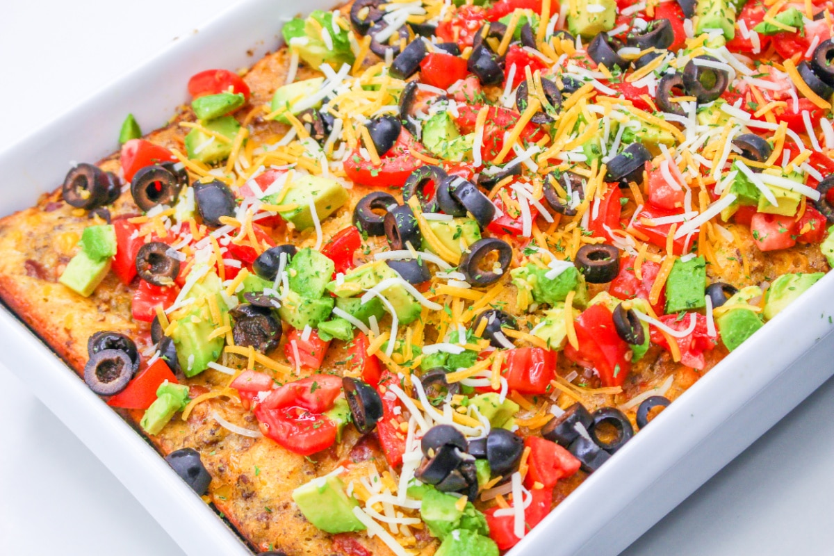 Cowboy Corn Casserole in a large pan topped with avocado, olives, and tomatoes.