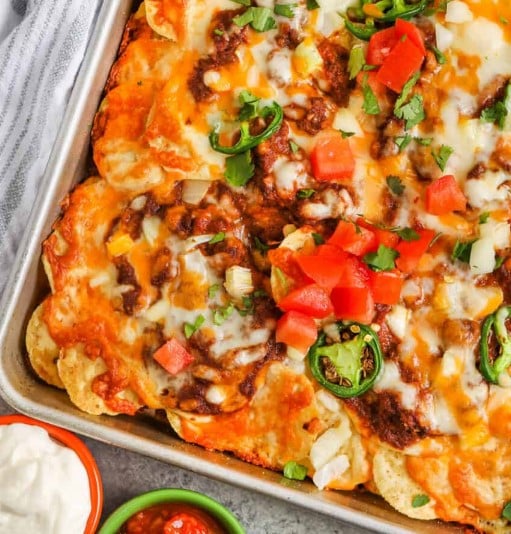 Chili Nachos on a sheet tray with chopped tomatoes and jalapenos on top.