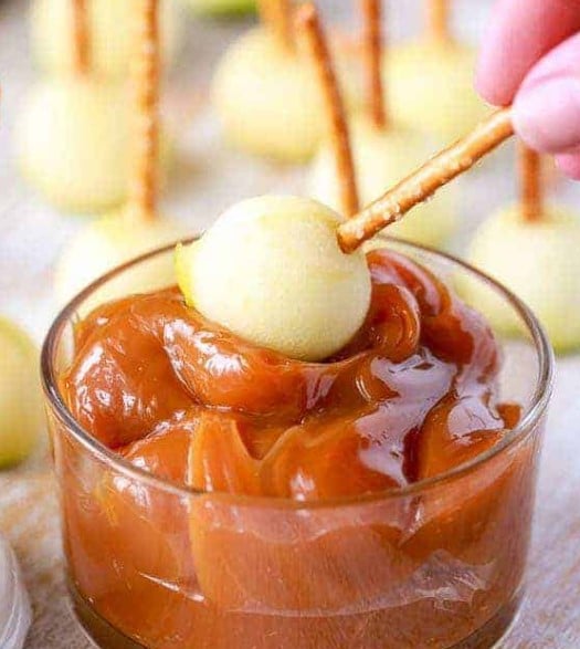 Hand holding a pretzel with a round piece of apple dipping into caramel.
